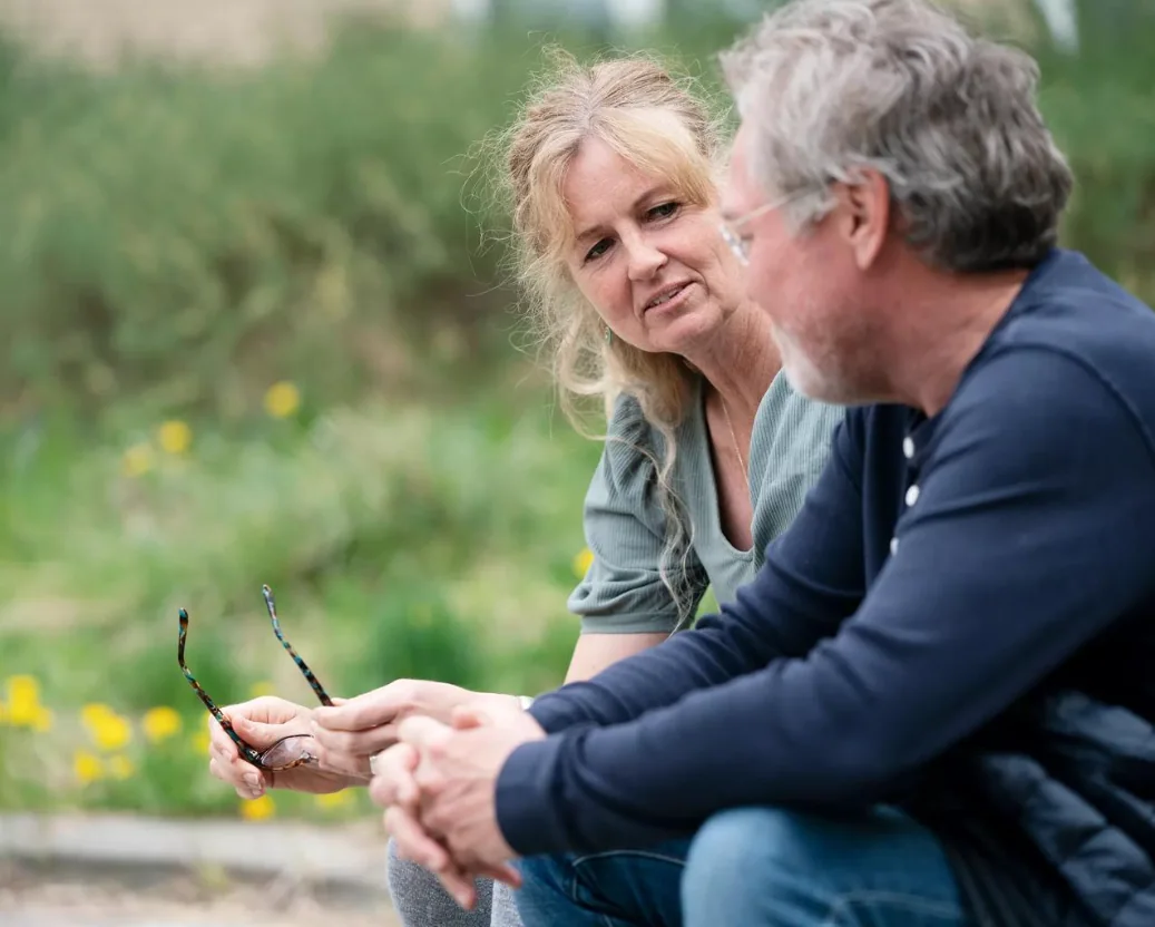 Two older adults talking together outdoors, surrounded by greenery.